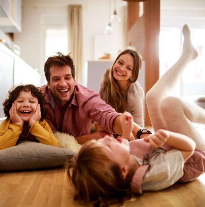 A joyful family laughing and playing on the floor, embodying the happy, healthy lives supported by Our Services at Renewal Family Dental.