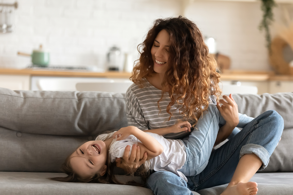 A joyful mother and daughter laughing and playing on a couch, embodying the happy families served by Renewal Family Dental.